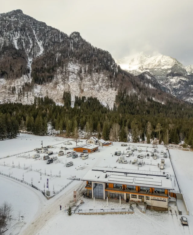 Luftaufnahme des verschneiten Campingplatzes mit Haupthaus vor Bergkulisse