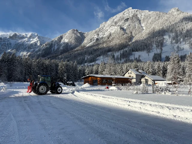 Traktor räumt die verschneite Straße am Campingplatz, im Hintergrund Berge und Holzhäuser