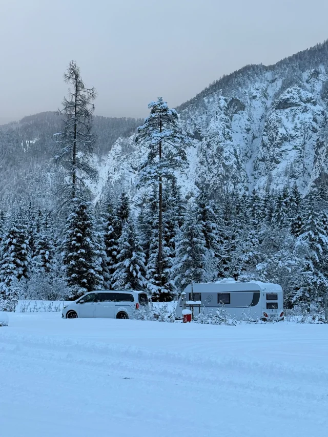 Pkw und Wohnwagen im verschneiten Tannenwald vor einem Berghang